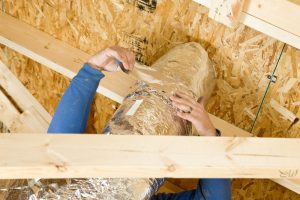 A worker is sealing two sections of house attic vent duct insulation with aluminum foil tape. The foreground is a section of truss and the background is OSB sheathing for the roof.